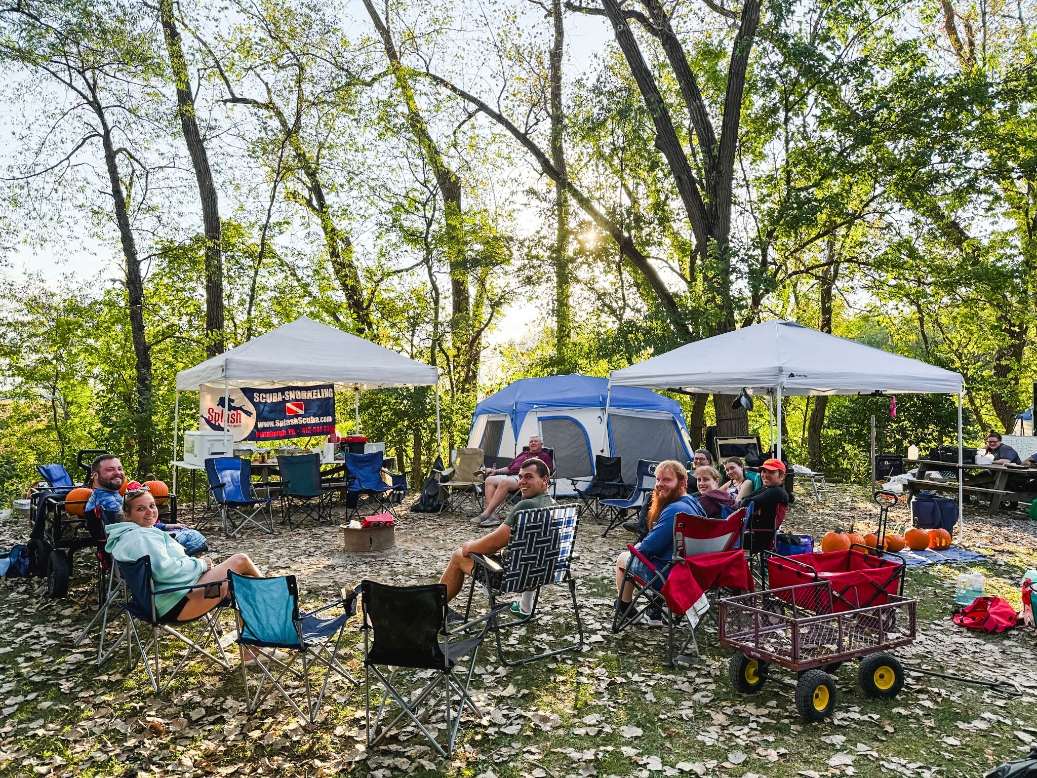 Group of campers relaxing in folding chairs around a wooded Gilboa Quarry campsite, with tents and pop‑up canopies set up, pumpkins and camping gear nearby, and sunlight filtering through the trees.